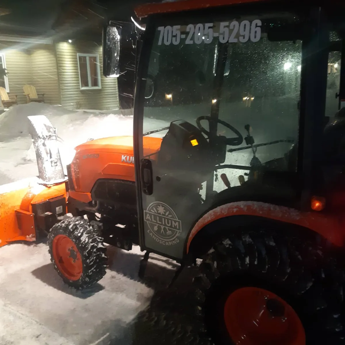An orange tractor with a snow blade beside a home at night